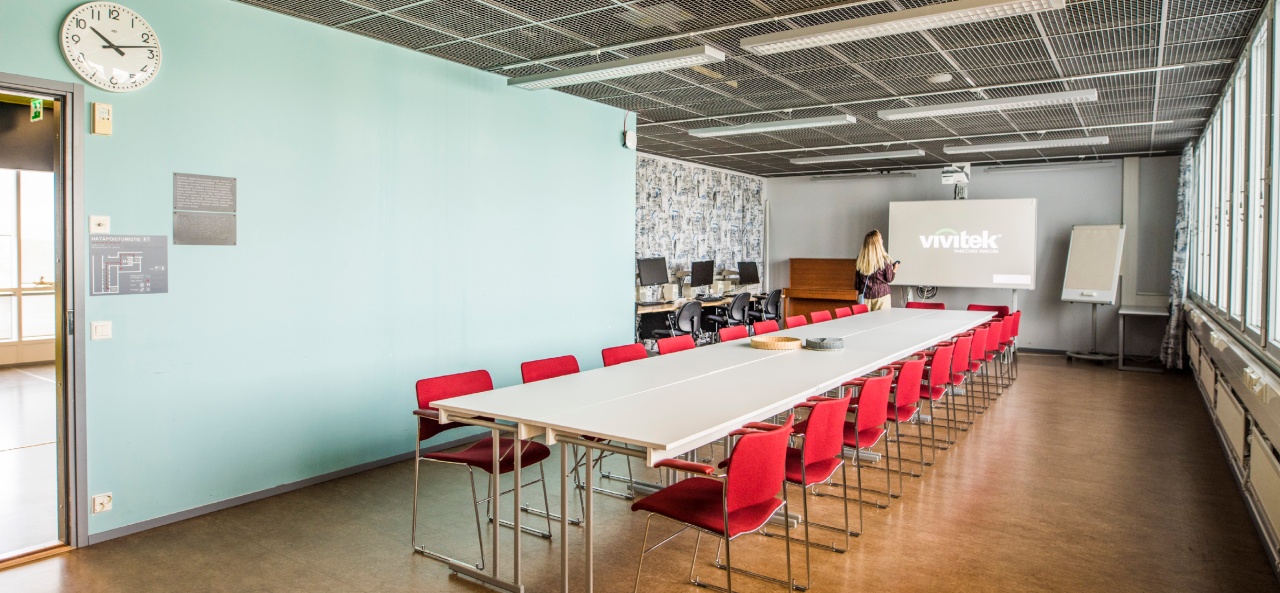 Pitkä, kapeahko tila, jonka keskellä pitkä vaalea pöytä, jonka ympärillä punaisia tuoleja. Pöydän päässä suuri näyttö. Takaseinustalla ruskea piano sekä pinottuja tuoleja.